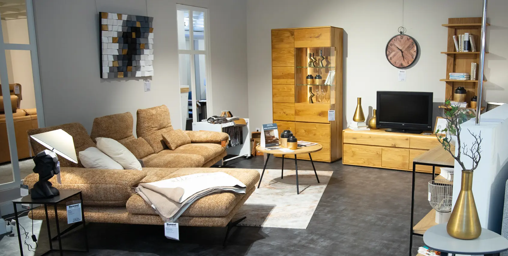 Sitzgruppe mit braunem Sofa, Couchtisch und Wohnwand aus Holzoptik, ergänzt durch Regal und Wandbild in neutralem Ausstellungsraum.